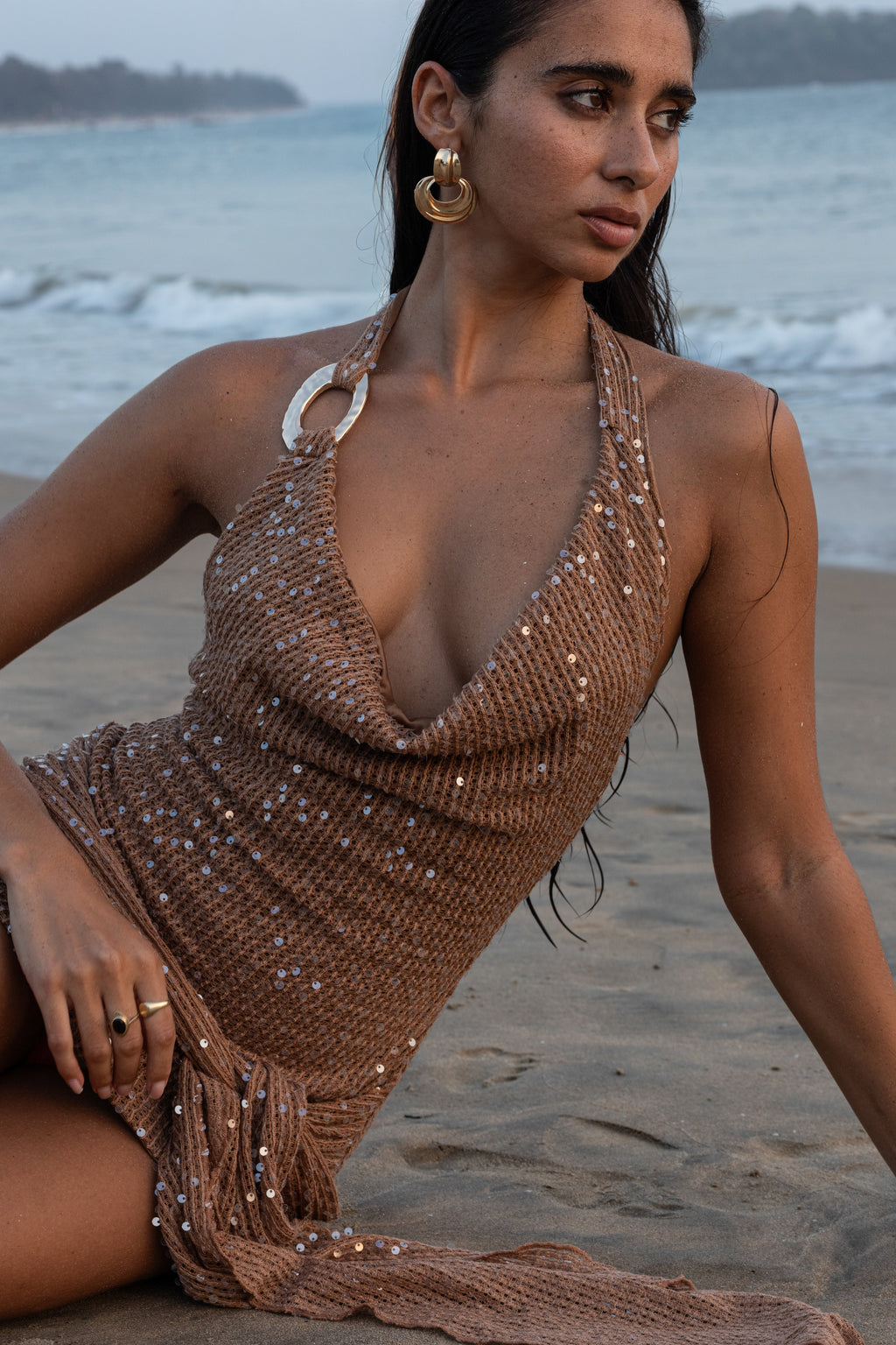 Woman in a brown sequin dress sitting on a beach with ocean in the background