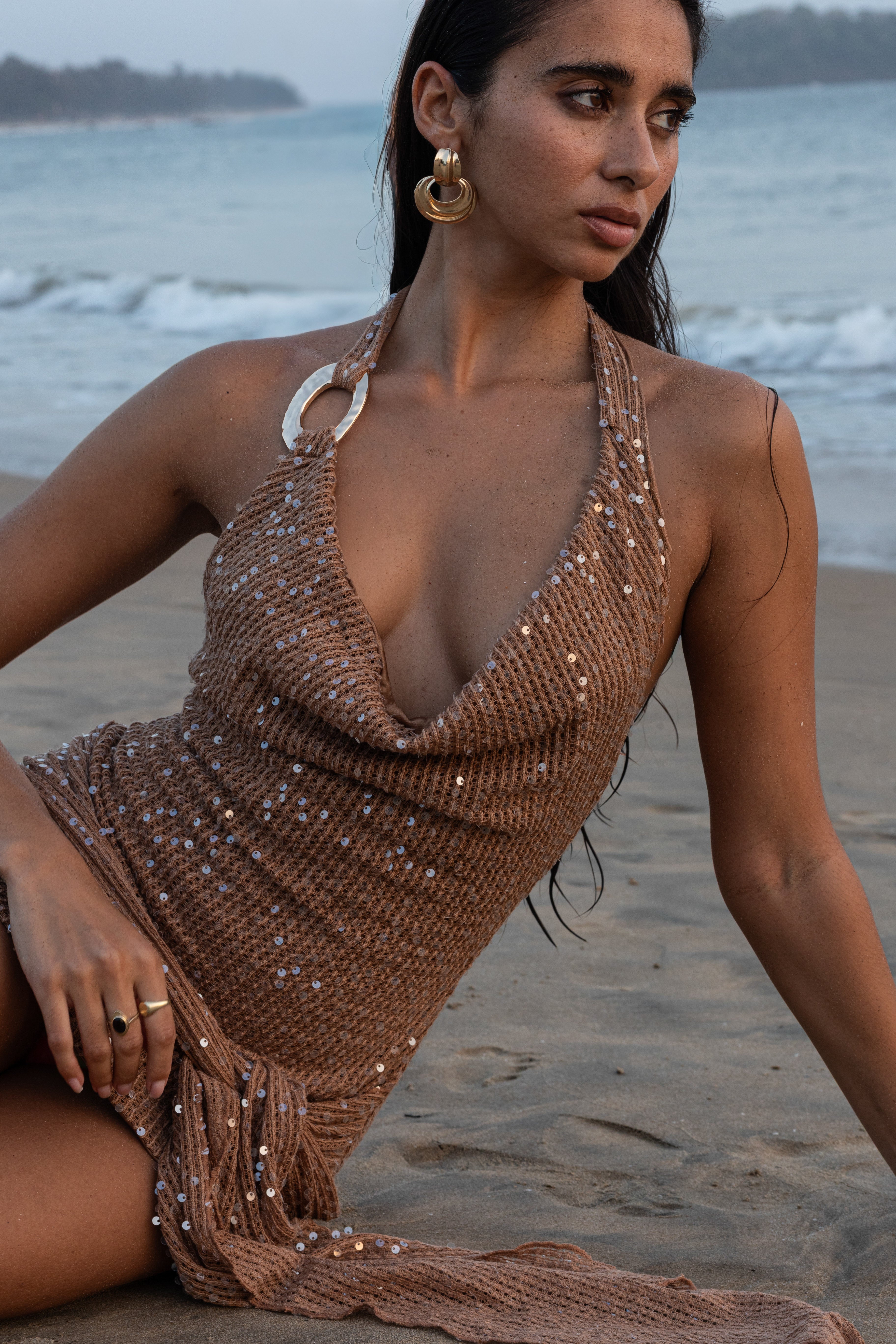 Woman in a brown sequin dress sitting on a beach with ocean in the background